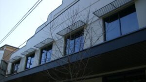 Modern slatted metal window awnings mounted above upper-floor glass windows on a building exterior.