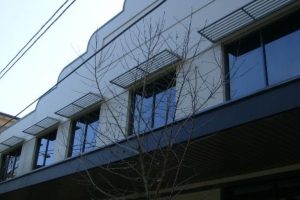 Modern slatted metal window awnings mounted above upper-floor glass windows on a building exterior.